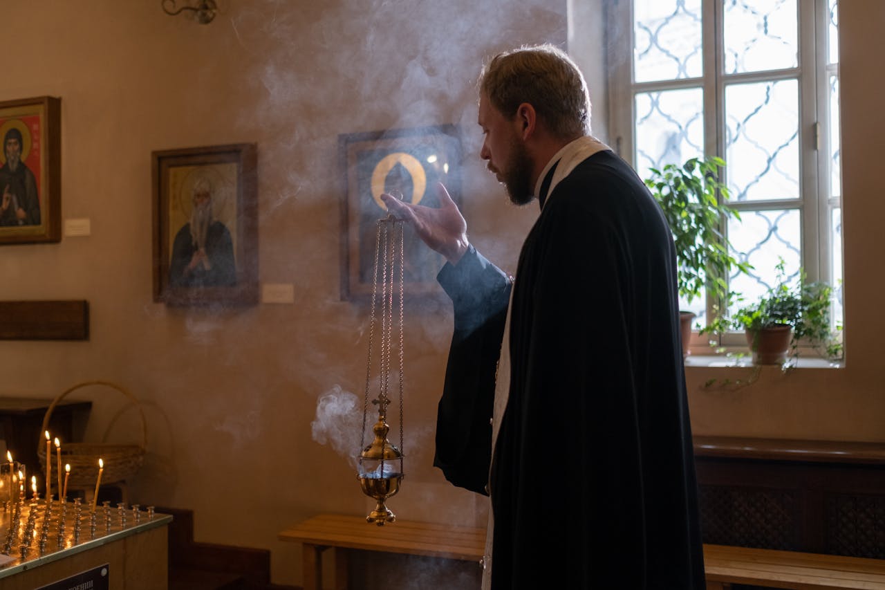 Orthodox priest holding censer in church, surrounded by religious icons and smoke.