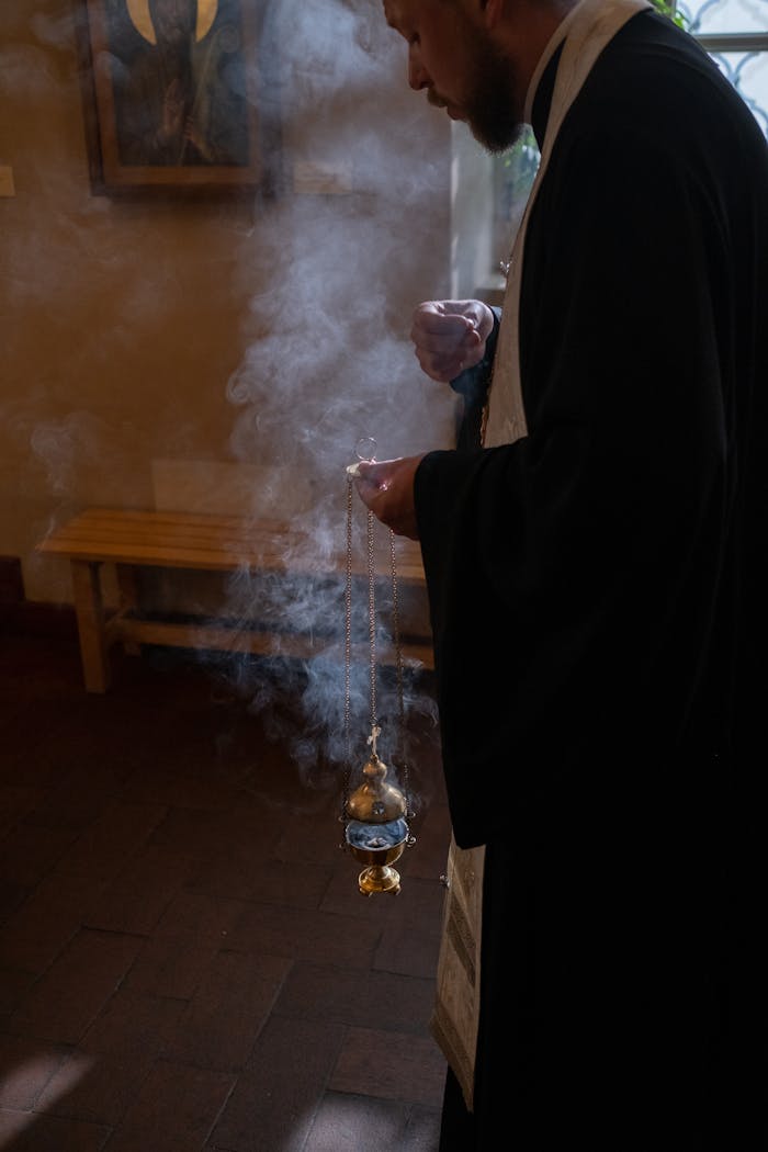 Orthodox priest holding incense during a religious ceremony, with smoke rising in a dimly lit church.